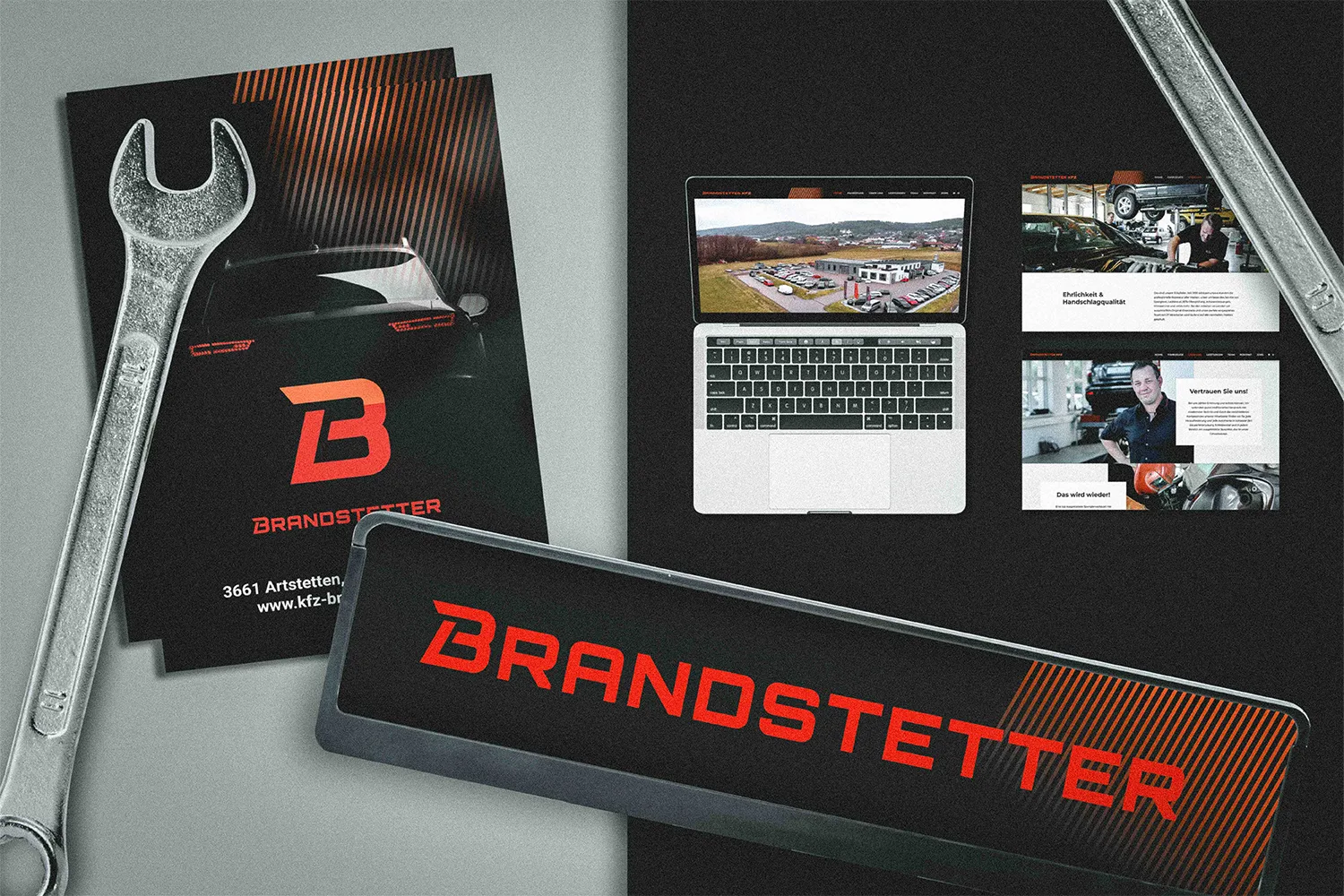 Gestaltetes Werbematerial für KFZ Brandstetter: schwarze Folder mit Sportwagen-Heck und Logo, offener Laptop mit Luftaufnahme des Firmenstandorts und Website-Ansichten mit Werkstatt und Mitarbeitenden, daneben ein Metallschild mit Brandstetter-Schriftzug und zwei großen Schraubenschlüsseln als gestalterisches Element.